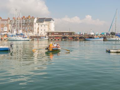 A harbor with people in a rowboat and moored sailboats with buildings and a pier in the background at The Oast House in Brewers Quay Harbour