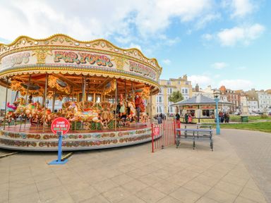 A carousel with people riding horses near a food kiosk and benches in a town square at The Oast House in Brewers Quay Harbour