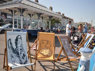 Deck chairs with various designs on a promenade with people and buildings in the background at The Old Boathouse in Weymouth