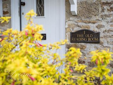 A white door with a decorative window and a plaque reading The Old Reading Room on a stone wall with yellow flowering plants in front at The Old Reading Room in Langton Herring