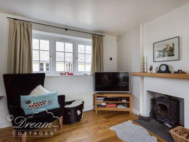 A living room with a black armchair, a wooden tv stand with a television, a fireplace with decorations, and a window with beige curtains at The Old Reading Room in Langton Herring