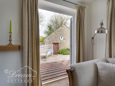 A living room with beige curtains a metal floor lamp and a sofa looking out to a patio and stone building at The Old Reading Room in Langton Herring
