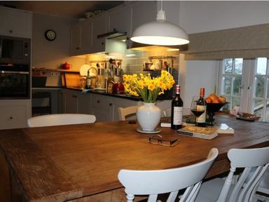 A kitchen with a wooden table holding flowers wine bottles books and glasses at The Old Reading Room in Langton Herring