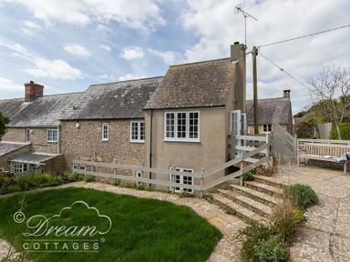 An exterior view of a stone and plaster cottage with a tiled roof a grassy area steps and a patio with seating at The Old Reading Room in Langton Herring