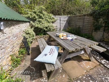 A wooden picnic table with cups plates and a cushion in a garden patio area at The Old Reading Room in Langton Herring