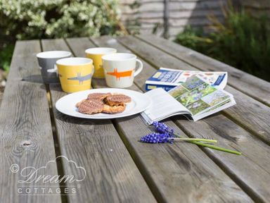 A wooden picnic table with four mugs a plate of biscuits an open book a flower bunch and a closed book at The Old Reading Room in Langton Herring