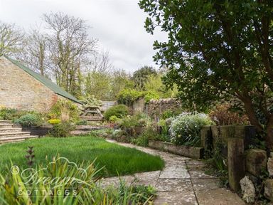 A garden with a stone path a grassy area plants trees wooden steps and a stone shed at The Old Reading Room in Langton Herring