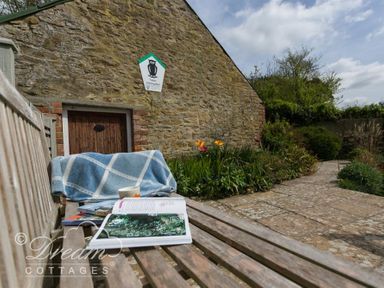 A wooden bench with a blue blanket a cup and an open book near a stone building and garden path at The Old Reading Room in Langton Herring