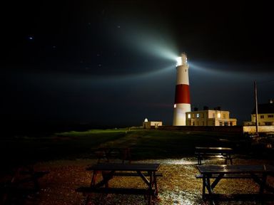 A lighthouse with beams of light illuminating the night sky near buildings and picnic tables at Olympic Cottage in Portland