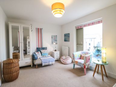 A room with a white wardrobe with mirrored doors a wicker basket a lounge chair with pillows and a throw a white nightstand with a lamp a bean bag a floral armchair by a window and a small round table with a lamp at Osprey House Portland