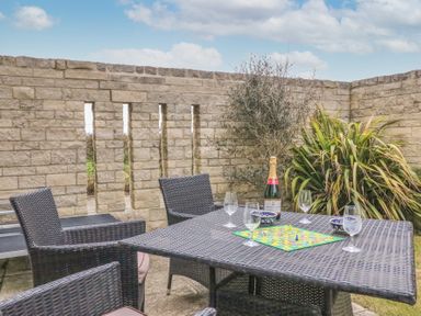 An outdoor patio with a wicker table and chairs a board game a bottle and glasses on the table and a stone wall with plants behind at Osprey House Portland