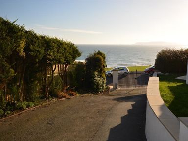 A driveway with bushes leading to a metal gate overlooking a road and the sea at Outlook Lodge in Bowleaze