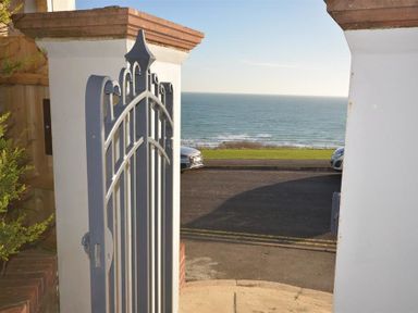 A metal gate opening to a view of a road grass and ocean at Outlook Lodge Bowleaze