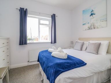 A bedroom with a bed covered by a blue blanket and two rolled towels a white chest of drawers and a window with dark curtains at Ocean Watch 1 in Greenhill