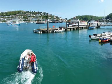 A person in a small motorized boat on water near a dock with several boats and houses on a hillside in the background at Bakers Dozen 13 Dartmouth House Dartmouth