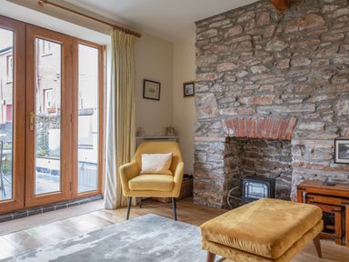 A sitting area with a yellow armchair and matching footstool next to a stone fireplace and wooden framed glass doors at 14a Courtenay Street Salcombe