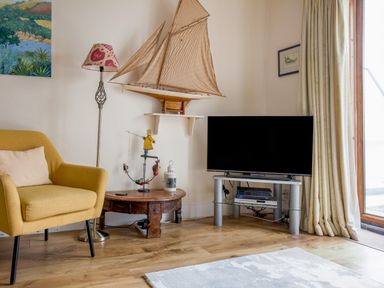 A living room with a yellow armchair a floor lamp a model sailboat on a wall shelf a wooden coffee table a television on a stand and curtains at 14a Courtenay Street Salcombe