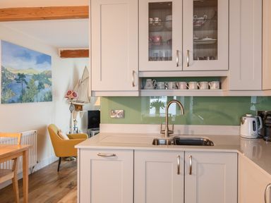 A kitchen with white cabinets a sink a kettle and a coffee machine with a green backsplash and a dining area with a yellow chair and wall art at 14a Courtenay Street in Salcombe