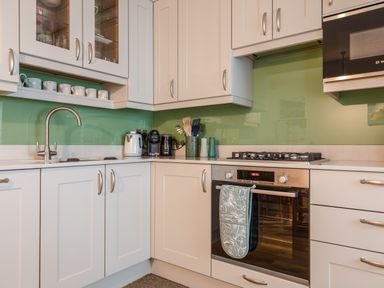 A kitchen with white cabinets, a green glass backsplash, a built-in oven, a gas stove, a kettle, a coffee machine, and mugs on a shelf at 14a Courtenay Street in Salcombe
