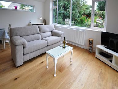 A living room with a grey sofa a glass top table with wine and glasses a television and wooden floor at Pebbles Burton Bradstock