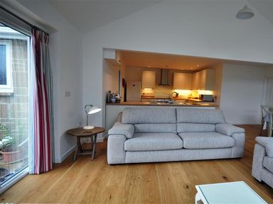 A living room with a gray sofa a wooden side table with a lamp and an open kitchen in the background at Pebbles in Burton Bradstock