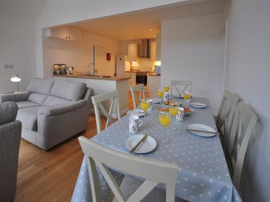 A dining table set with plates mugs and glasses of orange juice next to a living area with sofas and an open kitchen in Pebbles Burton Bradstock