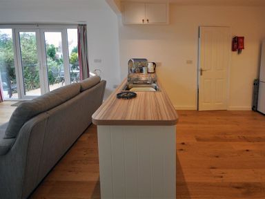 A kitchen island with sink and kettle next to a gray sofa and patio doors at Pebbles in Burton Bradstock