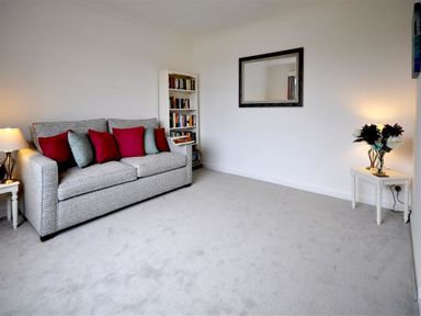 A living room with a grey sofa with red and green cushions a white bookshelf a mirror on the wall and a small table with a lamp and flowers at Pebbles in Burton Bradstock