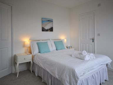 A bedroom with a double bed two bedside tables with lamps rolled towels on the bed and a beach painting on the wall at Pebbles in Burton Bradstock