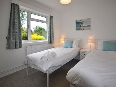 A bedroom with two single beds with white bedding and blue pillows with a window and nightstands with lamps at Pebbles in Burton Bradstock