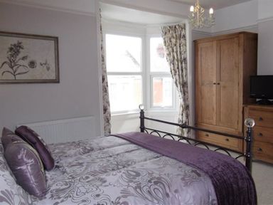 A bedroom with a patterned bedspread and pillows a wooden wardrobe and dresser and a window with curtains at Pedaller's Cottage in Weymouth