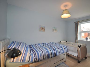 A bedroom with a bed with striped bedding a bedside lamp a portable crib and a window with curtains at Pedaller's Cottage in Weymouth