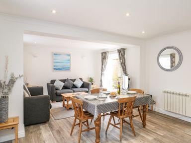 A dining area with a table and wooden chairs adjacent to a living room with sofas and a painting at The Pines in Weymouth