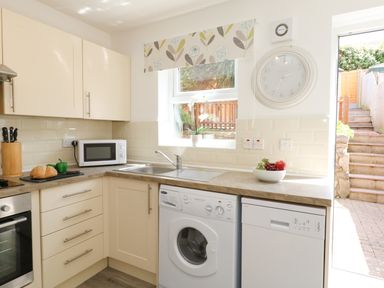 A kitchen with cream cabinets a washing machine a dishwasher a microwave and a window with floral blind at The Pines in Weymouth