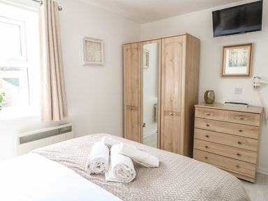 A bedroom with a bed, rolled towels on the bed, a wooden wardrobe with a mirror, a chest of drawers, a wall-mounted TV, and a window with beige curtains at The Pines in Weymouth