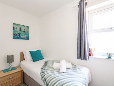 A single bed with white sheets and a green pillow next to a wooden nightstand with a lamp and a window with grey curtains and books on the sill at The Pines in Weymouth