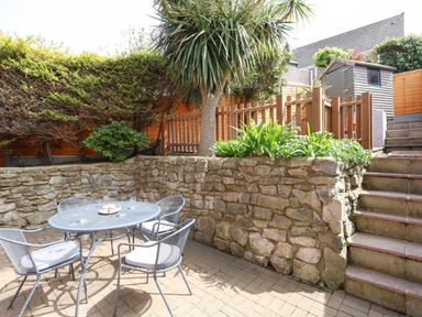 An outdoor patio area with a round metal table and four chairs next to stone walls plants and stairs leading to a wooden fence and shed at The Pines in Weymouth
