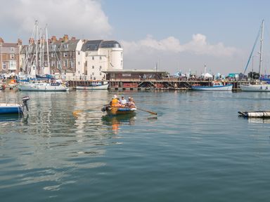 A small rowboat with four people on calm water near a quay with several moored sailboats and buildings at The Pines in Weymouth
