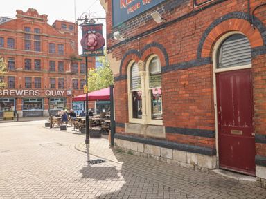 An outdoor seating area with people sitting at wooden tables near a brick building with a red door and The Red Lion sign and a large building with Brewers Quay sign in the background