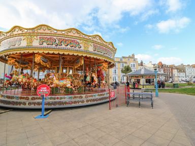 A carousel with painted horses and people riding it in an open area near buildings