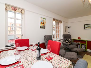 A dining area with a table set with wine glasses plates and a bottle of wine next to gray armchairs and a small green bookshelf at Poppies Brewers Quay Harbour