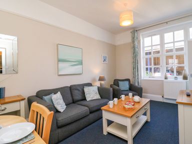 A living room with a dark sofa and armchair with cushions a wooden coffee table with mugs and games a window with curtains and a lamp at Reaver in Brewers Quay harbour