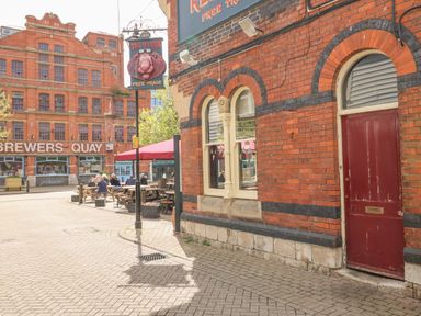 An outdoor seating area with people sitting at wooden picnic tables next to a red brick building with arched windows and a red door at Brewers Quay harbour