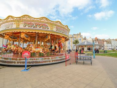 An outdoor carousel with people riding on painted horses near a kiosk and benches in a public square