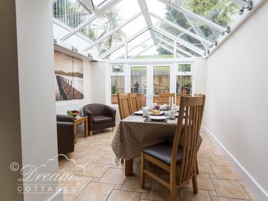 A dining area with a table set for a meal surrounded by wooden chairs and two armchairs under a glass roof at Redcliff View Lodge in Weymouth