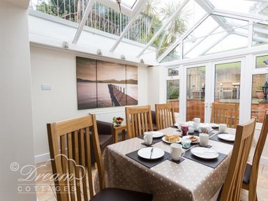 A dining area with a wooden table set for six under a glass roof with doors leading outside at Redcliff View Lodge in Weymouth