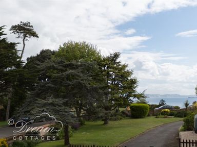 A garden with trees and bushes overlooking the sea with a driveway and parked cars at Redcliff View Lodge in Weymouth