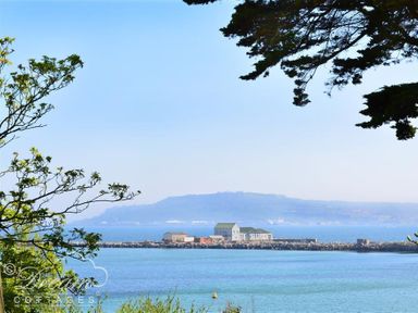 View of buildings on a pier with water and distant hills framed by tree branches at Redcliff View Lodge in Weymouth