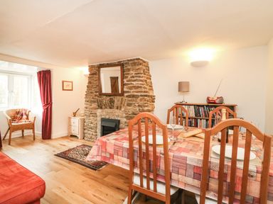 A dining area with wooden chairs and table set near a stone fireplace and a wicker chair by a window at River Cottage in Burton Bradstock