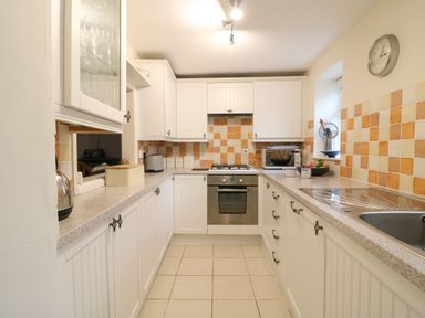 A kitchen with white cabinets orange and beige tile backsplash oven toaster microwave and a clock on the wall at River Cottage in Burton Bradstock